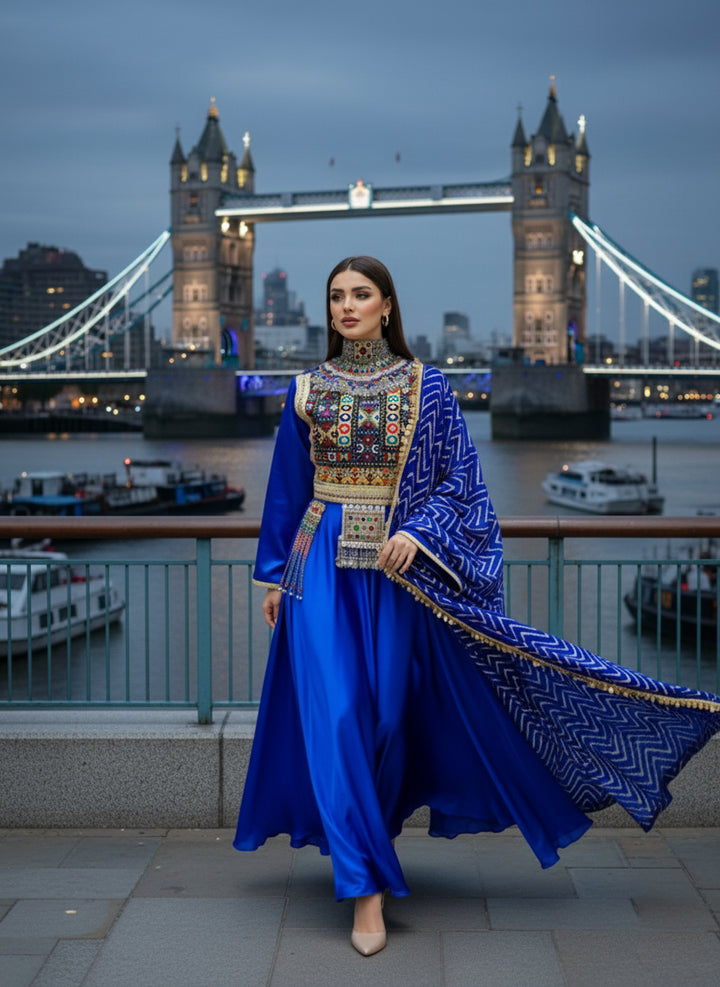 Woman in a blue traditional afghani outfit with intricate patterns standing in front of Tower Bridge at dusk.